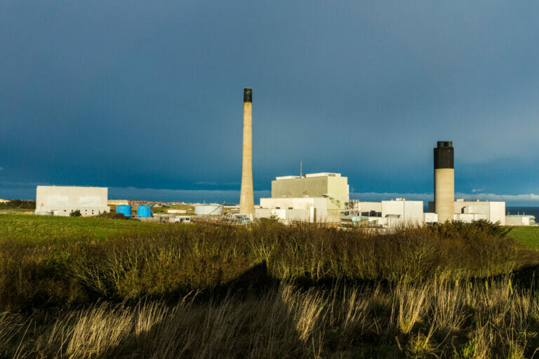 rticle headline: Plans to keep gas plant open could “blow hole” in climate targets Image description: Peterhead gas plant with tall chimneys and white industrial buildings sits under a dark, stormy sky in Aberdeenshire. Dry grass and bushes fill the foreground, with blue storage tanks and a glimpse of nearby town buildings beyond the station. Sunlight breaks through the clouds, casting a warm glow on the scene.