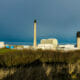 rticle headline: Plans to keep gas plant open could “blow hole” in climate targets Image description: Peterhead gas plant with tall chimneys and white industrial buildings sits under a dark, stormy sky in Aberdeenshire. Dry grass and bushes fill the foreground, with blue storage tanks and a glimpse of nearby town buildings beyond the station. Sunlight breaks through the clouds, casting a warm glow on the scene.