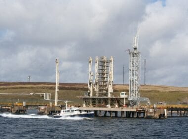 Article: "Surge in potent form of climate pollution at oil site" Image description: A large oil terminal jetty structure extending into the water, with pipes, cranes, and platforms visible for industrial operations. A small boat is moving past the jetty, leaving a wake in the water. The background shows rolling grassy hills and a partly cloudy sky.