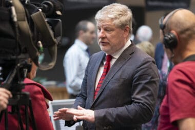 Article headline: When ‘critical friends’ fall out: how the Scottish Government sleepwalked into a crisis over meeting with an Israeli ambassador Image description: A man in a dark suit and red striped tie, speaking to journalists at a media event. He gestures with his hands while being filmed and recorded by a camera crew. Several people are blurred in the background.
