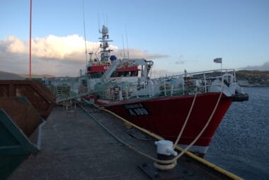 Article headline: Shetland fishing boat abandoned 80km of nets in seas for almost a month Image description: The red fishing vessel Genesis II (A100) moored at a pier, fishing nets draped over the side and coiled ropes securing the boat to the dock. A rugged coastal landscape is visible in the distance beneath a partly cloudy sky.