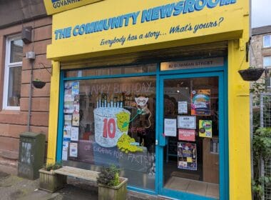article headline: Ten years on, The Ferret is still digging — and looking ahead alt text: A bright yellow shopfront with blue trim in Govanhill, Glasgow, reading “The Community Newsroom” and “Everybody has a story… What’s yours?”. The window is decorated with a large hand-drawn cake and the words “Happy Birthday to The Ferret — 10 years of digging for facts!”, celebrating The Ferret’s 10th anniversary. Posters and flyers fill the other windows, and a small bench with plants sits outside.