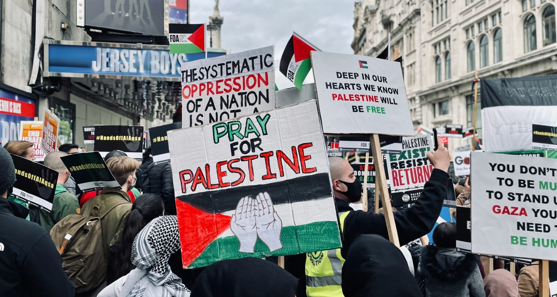 Article headline: Palestine Action: Why has the UK proscribed the group? Image description: People hold Palestinian flags and placards during a protest on a busy street in London’s West End, with theatre signs visible in the background.